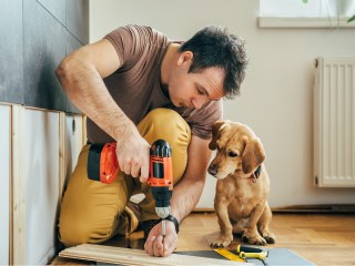 a man using a drill for home improvements while his dog sits beside him and watches him